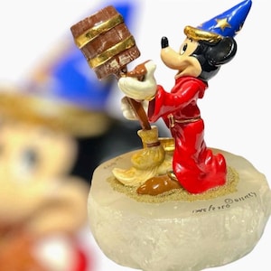 May include: A figurine of Mickey Mouse dressed as a sorcerer in a red robe and blue hat with gold stars, holding a brown wooden bucket and a broom. The character stands on a textured, translucent white base with the text Disney printed on it.