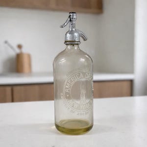 May include: Vintage glass seltzer bottle with a silver metal top. The bottle has a clear body with a circular embossed design that reads "America Dry New York". The bottle is filled with a light yellow liquid.