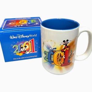 May include: A white ceramic mug with a blue interior and a white handle. The mug features the text "2001" in colorful letters with Mickey Mouse and other Disney characters. A blue box with "Walt Disney World" and "2001" is next to the mug.