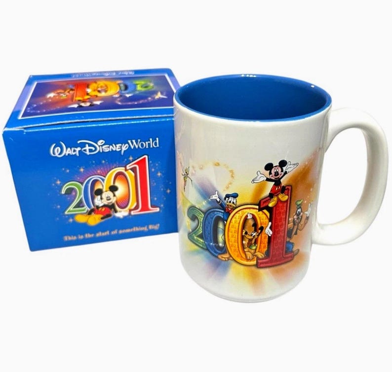 May include: A white ceramic mug with a blue interior and a white handle. The mug features the text "2001" in colorful letters with Mickey Mouse and other Disney characters. A blue box with "Walt Disney World" and "2001" is next to the mug.