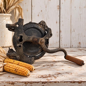 May include: A vintage cast iron hand-cranked corn sheller with a dark, weathered metal finish and a cylindrical wooden handle. The mechanical device features a circular central gear housing and a curved metal arm for manual operation.