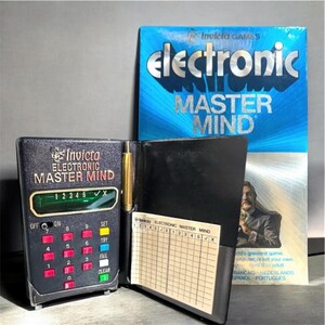 Puede incluir: Un juego Invicta Electronic Master Mind. El juego incluye una consola de juegos electrónica negra con botones numéricos rojos, un cuaderno negro con un bolígrafo y una caja azul con el título del juego.