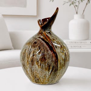May include: A ceramic vase with a rounded body and a flared, asymmetrical neck. The surface features a mottled glaze in shades of brown, tan, and cream, with a dark brown streak running vertically from the rim down the side of the vessel.