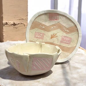 Może przedstawiać: Ceramiczna miska i pasujący okrągły talerz o kremowej podstawie z nakrapianą fakturą. Oba elementy są ozdobione wypukłymi, geometrycznymi wzorami w delikatnych odcieniach różu i beżu, w tym kwadratami, zygzakami i abstrakcyjnymi kształtami.