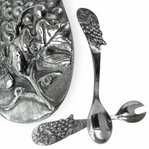 May include: Silver-toned serving utensils with grape cluster and leaf designs. The set includes a spoon, a fork, and a serving platter. The spoon and fork have curved handles. The platter has a detailed grape and leaf relief.
