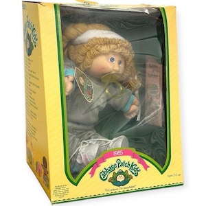 Puede incluir: Una muñeca Cabbage Patch Kids con cabello de lana rubio y una cinta blanca, vistiendo un chándal gris con detalles azules. La muñeca tiene un chupete en la boca y está empaquetada en una caja de cartón amarilla con el texto 1985.