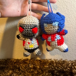 May include: Two crocheted toy figures hanging from keychains. The figure on the left is gray with red eyes and a yellow and blue chest. The figure on the right is blue with a red chest and white accents.
