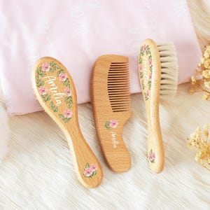 May include: A wooden baby grooming set featuring a brush, comb, and brush with soft bristles. Each item is decorated with pink roses and the name "Amelia" in script. The set is displayed on a soft pink blanket.