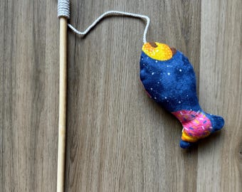 May include: A wooden fishing rod with a white string attached to a blue fabric fish toy. The fish has a colorful pattern with yellow, pink, and white accents. The toy is designed for children.