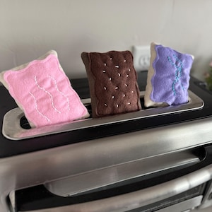Puede incluir: Una tostadora negra y plateada con tres pasteles de fieltro de colores en su interior. Los pasteles son rosas, marrones y morados, cada uno con detalles de costura blancos. La tostadora es de la marca Hamilton Beach.