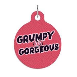 May include: A circular, pink pet tag featuring the text "GRUMPY BUT GORGEOUS" in black and white. The tag has a geometric design and a silver-coloured ring for attaching to a collar.