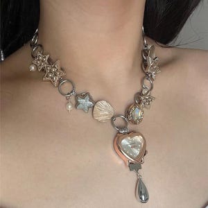 Peut inclure: Un collier argenté avec un pendentif montre en forme de cœur et divers breloques. Le collier présente des accents d'étoiles, de coquillages et de perles. La montre a un cadran blanc et un charme en forme de goutte d'eau argentée.