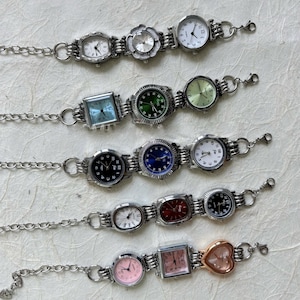 May include: A collection of silver-toned watch bracelets with various watch faces. The watch faces are in different shapes, including round, square, and heart-shaped, and feature different colored dials such as white, black, green, and pink.