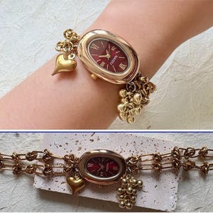 Pode incluir: Rel&oacute;gio de pulso dourado com mostrador oval bord&ocirc; e algarismos romanos. O rel&oacute;gio apresenta uma pulseira de elos e berloques decorativos, incluindo um cora&ccedil;&atilde;o e um grupo de pequenas esferas. O rel&oacute;gio tem aproximadamente 18 cm de comprimento.
