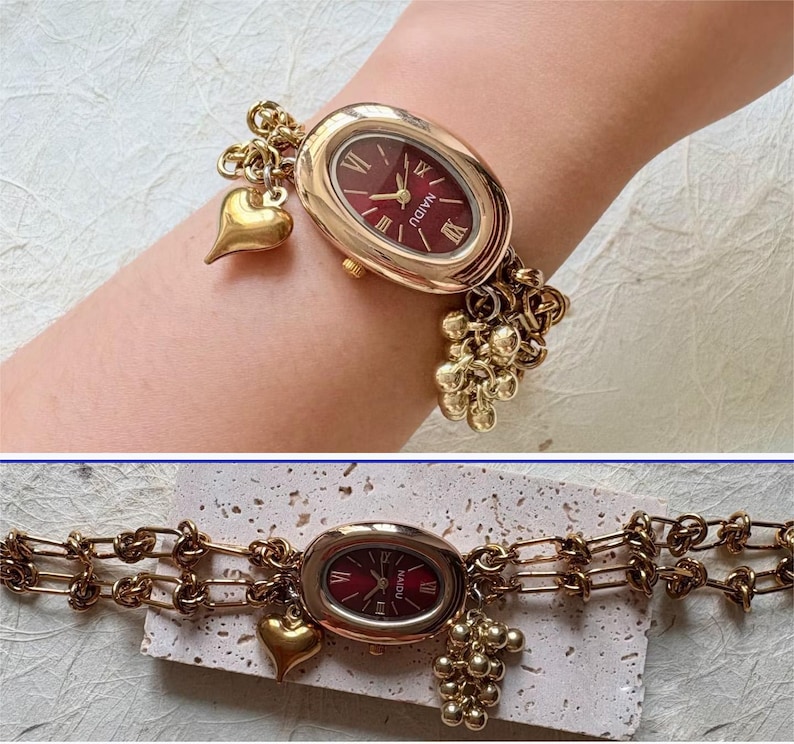 Pode incluir: Rel&oacute;gio de pulso dourado com mostrador oval bord&ocirc; e algarismos romanos. O rel&oacute;gio apresenta uma pulseira de elos e berloques decorativos, incluindo um cora&ccedil;&atilde;o e um grupo de pequenas esferas. O rel&oacute;gio tem aproximadamente 18 cm de comprimento.