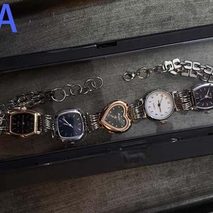 May include: A collection of silver and gold-toned watches with various face shapes, including rectangular, square, and heart-shaped, displayed in a black case. The watches have metal link bands and different colored watch faces.