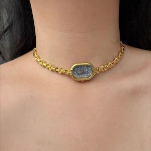 May include: A gold-toned watch necklace with an oval watch face and a linked chain. The watch face has a blue background with gold-colored hands and markers. The necklace is worn around the neck.