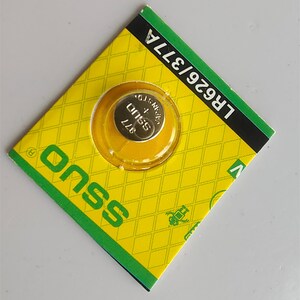 Pode incluir: Uma pequena pilha prateada 377 &eacute; vis&iacute;vel atrav&eacute;s de uma janela de pl&aacute;stico transparente numa embalagem amarela e verde. A embalagem tem um padr&atilde;o de losangos e a marca "onss" est&aacute; impressa na frente. O texto "LR626/377A" est&aacute; impresso.
