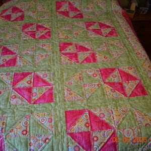 May include: A quilted bedspread featuring a repeating block pattern of solid hot pink fabric and a multi-colored floral print on a light sage green background. The quilt has a visible stitched texture and includes a date stamp in the bottom right corner.