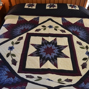 Op de afbeelding: Een quilt met een grote, witte ster in het midden, omringd door een donkerblauwe en bordeauxrode rand. De ster is gemaakt van kleinere sterren en is omgeven door een bloemmotief. De quilt is gemaakt van katoenen stof en is in uitstekende staat.