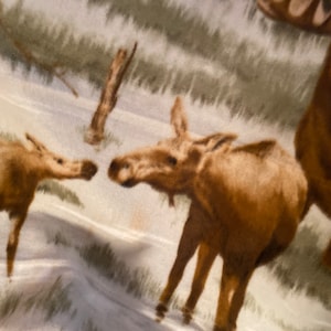 Puede incluir: Una tela blanca con un alce marrón y un cervatillo impresos en ella. El alce y el cervatillo están de pie en un campo de hierba.