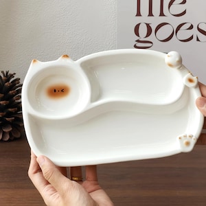 May include: White ceramic cat-shaped divided plate. The plate features a round bowl with a brown face design and two divided sections. The plate has cat ears, a paw, and a tail design. The plate is held by hands.