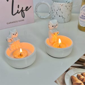 以下が含まれることがあります： 猫の形をした白いセラミック製のキャンドルホルダーが2つ。それぞれに小さなティーライトキャンドルが入っており、炎が灯っています。キャンドルホルダーは白い表面の上に置かれています。