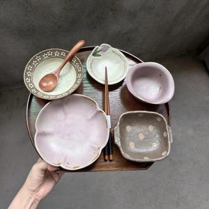 Puede incluir: Una bandeja de madera oscura contiene una colección de vajilla de cerámica que incluye un cuenco redondo con borde floral y cuchara de madera, un plato pequeño en forma de pétalo, un cuenco rosa liso, un cuenco cuadrado con lunares y palillos.