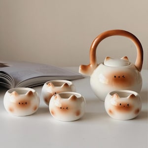 Op de afbeelding: Een keramische theeservies met een kattengezichtontwerp, bestaande uit een theepot met een bruine handgreep en tuit, en vier kopjes. De set is wit met bruine details en staat op een wit oppervlak.