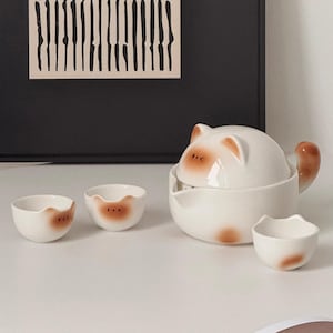 May include: A white ceramic cat-themed tea set, including a teapot, lid, and three small cups. The teapot features a tail-shaped handle and brown detailing on the ears and face. The cups also have brown accents. The set is on a white surface.