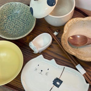 May include: A collection of ceramic tableware, including bowls, a cat-shaped plate, and a small dish. The bowls feature various designs and colors, with a wooden spoon and chopsticks also present. The items are arranged on a wooden tray.