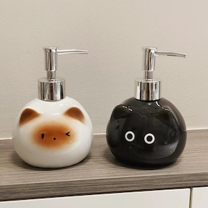 Op de afbeelding: Twee keramische zeepdispensers. Eén is wit met bruine beerachtige kenmerken, de andere is zwart met witte ogen. Beide hebben zilveren pompknoppen en staan op een houten plank.