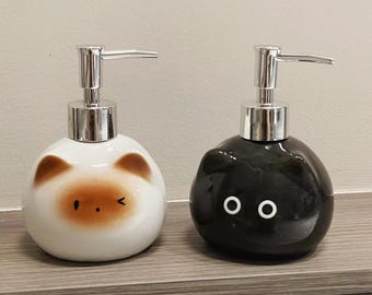 Può includere: Due dispenser di sapone in ceramica. Uno è bianco con caratteristiche simili a quelle di un orso marrone, l'altro è nero con occhi bianchi. Entrambi hanno pompe argentate e sono su una mensola di legno.