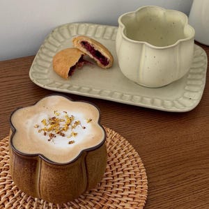 May include: A brown, flower-shaped ceramic mug filled with a latte topped with dried yellow flowers, sitting on a woven mat. Beside it, a rectangular, scalloped-edge ceramic tray holds a light-colored empty mug and a halved berry pastry.
