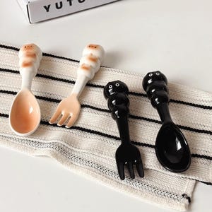 May include: Four ceramic spoons and forks with cartoon character handles. Two are white with tan accents, and two are black. The spoons and forks are arranged on a white and black striped cloth.