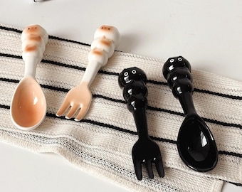 May include: Four ceramic spoons and forks with cartoon character handles. Two are white with tan accents, and two are black. The spoons and forks are arranged on a white and black striped cloth.
