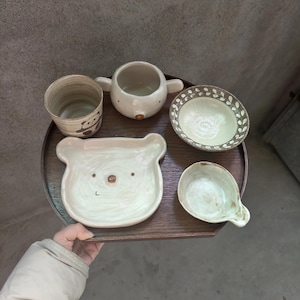 May include: A collection of five ceramic pieces on a dark brown wooden tray: a cylindrical cup with a bear illustration, a small round bowl with handles, a shallow bowl with a leaf-patterned rim, a bear-shaped plate, and a small pouring dish.