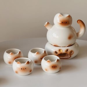Könnte beinhalten: Ein weißes Keramik-Teeservice mit Katzenmotiv. Das Set umfasst eine Teekanne, einen Wärmer und vier kleine Teetassen. Die Teekanne hat einen katzenförmigen Deckel und Griff, und der Wärmer hat ein Pfotenabdruck-Design. Die Teetassen haben Katzengesichter.
