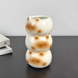 Op de afbeelding: Een gestapelde keramische plantenbak in de vorm van drie kattenkoppen. De witte plantenbak heeft bruine vlekken en zwarte ogen, met een ronde opening aan de bovenkant. Het gestapelde ontwerp creëert een grillig en decoratief stuk.
