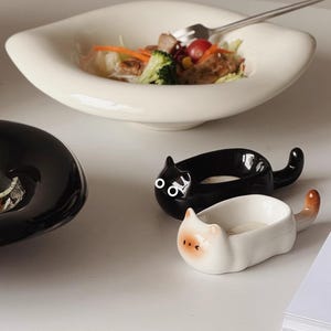 Puede incluir: Un cuenco de cerámica blanca con ensalada y un tenedor. También hay dos cuencos de cerámica con forma de gato, uno negro y otro blanco. El cuenco de gato blanco tiene una nariz y una cola de color melocotón.