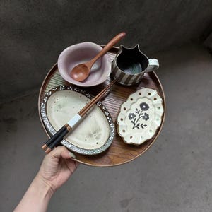 May include: A round wooden tray holding a pink ceramic bowl with a wooden spoon, a striped ceramic mug, a pair of wooden chopsticks with white and black accents, an oval plate with a floral border, and a small scalloped plate with a black floral design.