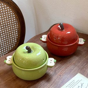 May include: Two ceramic lidded bowls shaped like apples, one in bright red and one in light green. Each bowl features a dark brown stem on the lid and two small, flat, white apple-shaped handles on the sides with green leaf accents.
