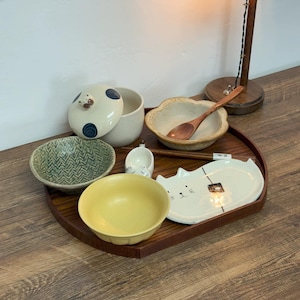 May include: A collection of ceramic tableware including bowls, a lidded container, and a cat-shaped plate. The set features a variety of colors and patterns, including a yellow bowl, a green patterned bowl, and a wooden spoon. The items are arranged on a wooden tray.