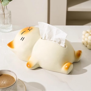 May include: A cream-coloured ceramic tissue box in the shape of a cartoon cat, with orange accents on the ears and paws. The cat is lying down, with a slot on its back for tissue dispensing. White tissues are visible.