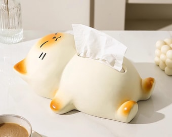 Puede incluir: Un dispensador de pañuelos de cerámica color crema con forma de gato de dibujos animados, con detalles en naranja en las orejas y las patas. El gato está tumbado, con una ranura en la espalda para dispensar pañuelos. Se ven pañuelos blancos.