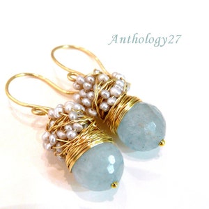 May include: Gold earrings with a light blue gemstone and white pearl accents. The gemstone is faceted and the pearls are wrapped in gold wire.