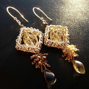 May include: Gold tone earrings with a square-shaped cage design featuring a clear, faceted gemstone. The earrings have a dangling teardrop-shaped gemstone and brown beads.