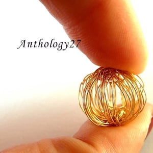 May include: A small, spherical, gold-coloured wire sculpture held between two fingers. The wire is intricately woven, creating a cage-like design. The text "Anthology27" is visible in the upper left corner.