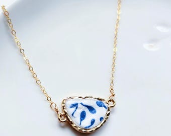 Könnte beinhalten: Eine goldfarbene Halskette mit Anhänger. Der Anhänger hat einen weißen Hintergrund mit blauem Blumenmuster, eingefasst von einer goldfarbenen Lünette. Die Halskette wird auf einer weißen Oberfläche präsentiert.