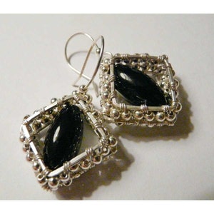 May include: Silver earrings with black gemstone accents. The earrings feature a square frame with a black oval gemstone in the center. The frame is made of silver wire and beads.
